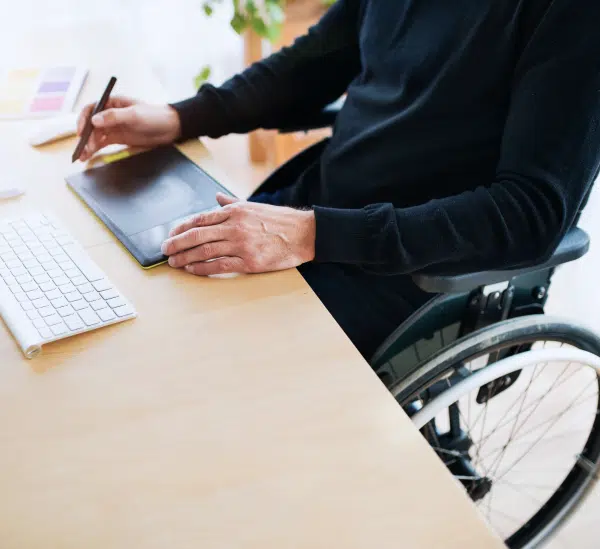 Person in a wheelchair working at a desk with a tablet and keyboard, representing workplace inclusion for individuals with disabilities.
