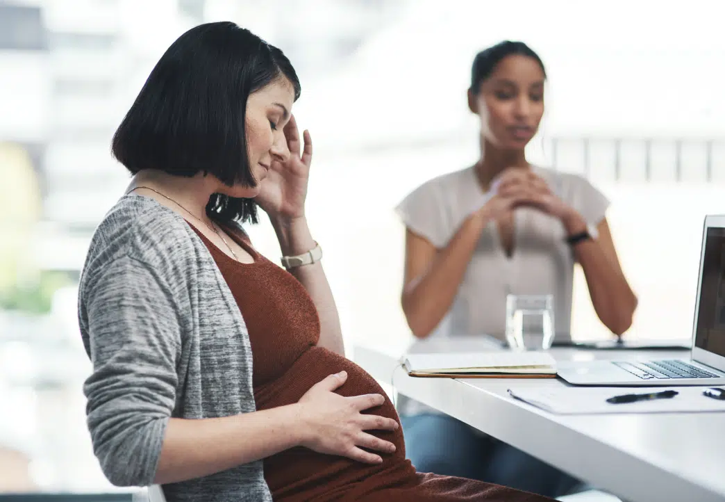 Pregnant woman in distress sitting at a table during a consultation, holding her belly with one hand and forehead with the other.








